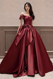 Woman wearing a burgundy off-shoulder gown with a large bow detail against a neutral wall.