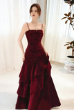 Woman wearing a burgundy evening gown indoors with white curtains and a plant in the background.