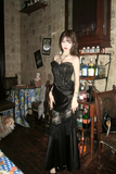 Woman in a black dress standing in a room with vintage decor and bottles on shelves.