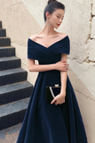 Woman in a navy blue off-shoulder dress holding a clutch against a textured wall.