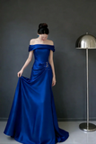 Woman in a blue off-shoulder gown standing against a dark gray wall with a floor lamp.