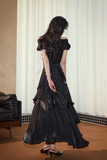 Woman wearing a black off-shoulder dress in a room with curtains and a chair.