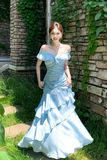 A woman wearing a light blue off-the-shoulder mermaid-style gown stands outdoors against a stone wall with green ivy and grass.