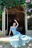 A woman wearing a light blue off-the-shoulder gown poses on a stone ledge in front of open white doors, with lush greenery, purple flowers, and a chandelier visible in the background.