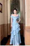 A woman stands in an indoor hallway wearing a light blue off-the-shoulder gown with a fitted bodice, bow detail, ruching, and a layered ruffled skirt.