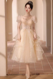 A woman wearing a pale champagne-colored tulle dress with puff sleeves, a sweetheart neckline, and a layered ruffled skirt, standing in a softly lit arched hallway.