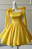 A bright yellow satin mini dress with long draped sleeves and large bow details on the shoulders, displayed on a white mannequin against a light gray paneled wall.