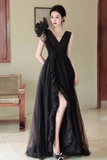 A woman models a floor-length black evening gown with a high slit, ruffled layered skirt, and a large black feather embellishment on the shoulder, set against an indoor background.