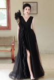 A woman models a long black evening gown with a V-neckline, ruffled sleeve detail, and a high leg slit, standing in an indoor setting with a bench and window in the background.