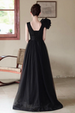 A woman wearing a long black evening gown, photographed from the back in an indoor setting with a window and chair visible in the background.