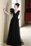 A woman wearing a floor-length black formal gown with a deep V-neckline, sheer tulle overlay, and a large ruffled shoulder detail, posed indoors against a light wall near a window.