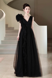 A woman wearing a floor-length black evening gown with a ruffled one-shoulder design and sheer layered skirt, standing in front of a modern curved staircase.