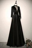 A floor-length black evening gown displayed on a mannequin against a plain light wall. The dress has a sheer black high-neck overlay, a deep V-neckline, three-quarter sleeves, a fitted waist, and a full flowing skirt.