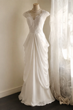 A white wedding dress displayed on a mannequin in an indoor setting, featuring a lace bodice, cap sleeves, and a draped satin skirt, with sheer curtains to the right.