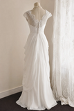 A white wedding gown displayed on a dress form mannequin, featuring a lace V-neck bodice, cap sleeves, and a flowing chiffon skirt, set against a neutral indoor background with a sheer curtain.