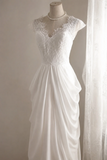 A white wedding dress displayed on a mannequin, featuring a lace bodice with cap sleeves, a V-neckline, and a flowing draped skirt, set against a neutral background with sheer curtains.