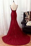 A deep burgundy red lace mermaid evening gown displayed on a mannequin, featuring a low open back, spaghetti straps, and a long train on a wooden floor next to a black leather armchair.