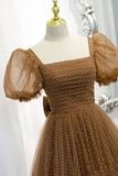 A brown tulle dress with white polka dots displayed on a cream mannequin dress form with a gold neck cap. The dress features a square neckline, voluminous puff sleeves with fringe, and a gathered skirt.