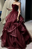 A woman wearing a strapless ball gown in deep burgundy with a sweetheart neckline, ruched bodice, and a voluminous tiered tulle skirt with subtle sparkle, standing indoors.