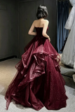 A woman stands with her back to the camera wearing a strapless ball gown in deep burgundy with sparkly embellishments and layered ruffles.