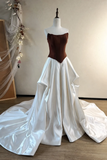 A strapless formal gown displayed on a mannequin in an indoor studio-like setting. The dress has a fitted dark brown bodice and a voluminous white satin skirt with a long train.