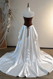 A white satin ball gown with a structured brown corset-style bodice and long tie, displayed on a dress form mannequin in an indoor room with a beige wall and wood floor.