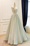 A sleeveless ball gown in a pale sage green or mint green shade, displayed on a mannequin in an indoor room with white paneled walls, a dark blue curtain, and a patterned rug.