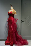 A formal evening gown displayed on a mannequin indoors. The dress is predominantly bright red with dense sequin embellishment, a sheer off-white lace upper bodice, and gold floral applique.