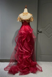 A formal ball gown displayed on a mannequin in an indoor room. The dress has a deep red ruffled tulle skirt, a red beaded bodice, and gold floral lace off-the-shoulder sleeves with a matching gold collar.