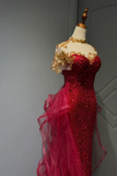 A red sequined evening gown displayed on a cream dress form with a gold neck cap. The dress features gold floral lace applique at the off-the-shoulder neckline, a sweetheart neckline, and a layered, sheer magenta tulle train.