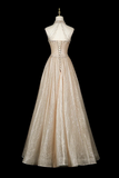 A floor-length formal gown displayed on a mannequin against a solid black background. The dress is champagne beige with a shimmering, textured tulle skirt and a lace-up corset-style bodice with crystal embellishments.