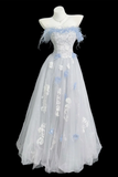 A formal off-the-shoulder gown displayed on a white mannequin against a black background. The dress features a fitted lace bodice with light blue feather accents, a matching waistband, and a voluminous tulle skirt decorated with white floral appliqués and light blue flower embellishments.