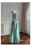A floor-length satin slip dress in a soft mint green color, displayed on a white mannequin in a bright room with a wooden floor, curtains, and a fireplace.