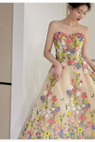 A woman wearing a strapless sweetheart ball gown with a beige tulle base and colorful floral embroidery in pink, yellow, blue, and white, standing indoors.