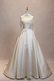 An off-white satin ball gown displayed on a mannequin against a neutral beige backdrop. The dress features a structured, draped bodice with a wide shoulder yoke and a voluminous, flowing skirt.