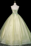 A strapless ball gown displayed on a mannequin against a black background. The dress is a pale, shimmering yellow-green with a structured corset-style bodice, floral applique details, and a voluminous tulle skirt.