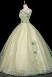 A strapless ball gown displayed on a mannequin against a black background. The dress is a pale yellow-green tulle with floral appliques in matching green and white, featuring a sweetheart neckline and a voluminous skirt.