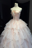 A pale blush pink strapless ball gown with a large bow at the bust and a layered tulle skirt, displayed on a white mannequin against a dark background.