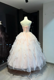 A pale blush pink ball gown displayed on a mannequin in a showroom. The strapless gown features a structured bodice with a central bow and a voluminous, multi-layered ruffled tulle skirt.