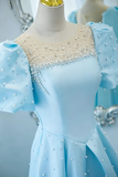 A light blue formal dress displayed on a cream mannequin torso. The dress features a sheer illusion neckline embellished with white pearls and crystal beadwork, short puff sleeves with matching pearl details, and a flared skirt.