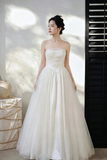 A woman wearing a strapless white ball gown with a fitted bodice and voluminous tulle skirt, standing indoors against a sunlit wall with shadows.