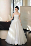 A woman wearing a strapless white ball gown with a fitted bodice and voluminous tulle skirt, standing indoors near large windows and neutral-toned architectural elements.