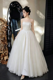 A woman wearing a strapless white ball gown with a full tulle skirt, standing in a styled indoor setting with a large black sculptural mirror and dried botanical decor.