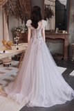 A woman stands with her back to the camera wearing a voluminous tulle ball gown with floral lace applique and a corset-style bodice, photographed in a rustic indoor room with wood furniture and dried floral decor.