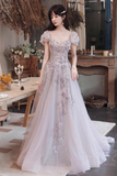 A woman models a formal A-line gown with short puff sleeves, a square neckline, and intricate floral embroidery and beadwork on a light lavender-gray tulle skirt, set in a vintage-style indoor room.