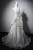 A strapless white wedding gown with a ruched bodice and dramatic draped overskirt, displayed on a white mannequin against a dark gray studio background.