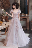 A woman wearing a pale pink tulle ball gown with floral lace applique and puff sleeves, standing in a rustic indoor room with a table and dried flowers in the background.